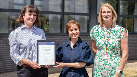Three people standing outdoors in front of a building with large windows; one holds a framed certificate